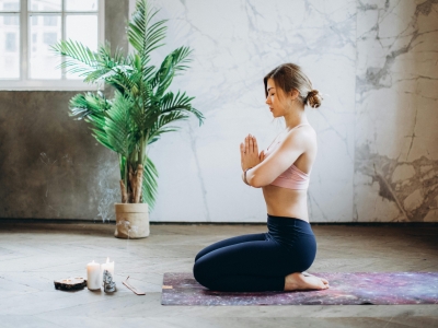 Eine junge Frau, bekleidet mit einem rosa Oberteil und einer schwarzen Hose kniet auf einer bunten Yogamatte.