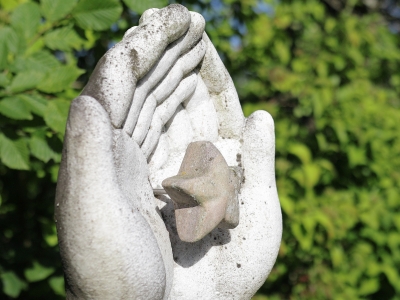 Zwei Hände aus Stein, die in ihrer Mitte einen braunen Stern halten. Im Hintergrund sind grüne Hecken zu sehen, die von der Sonne angestrahlt werden.